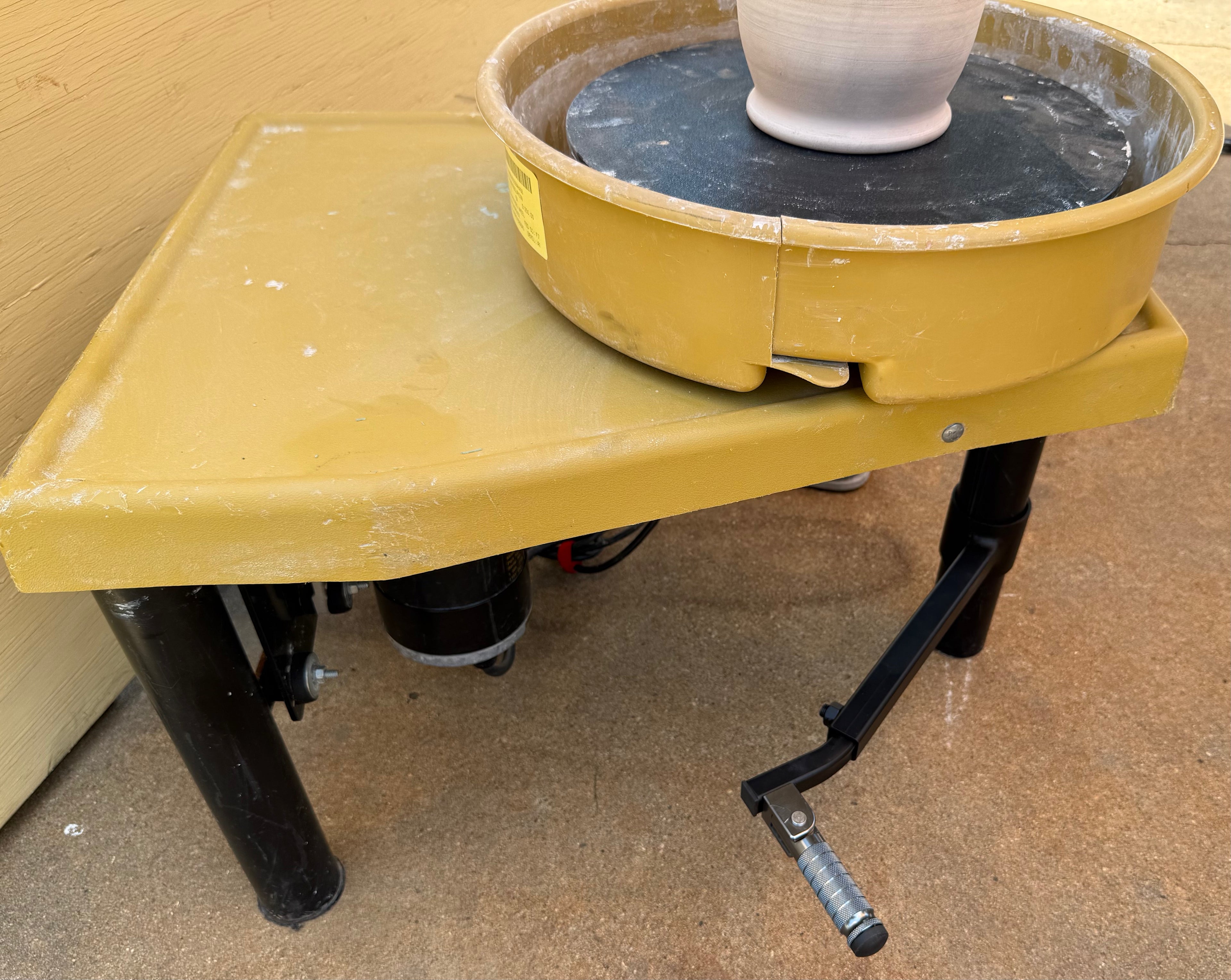 A yellow electric pottery wheel stand with The Potters Peg, a black crank handle, shown with a pottery wheel and a white ceramic pot on top, set against a tan wall and concrete floor.